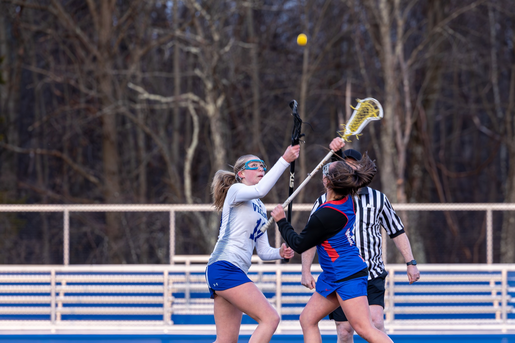 Two girls playing lacrosse on the field with the ball in the air above them.
