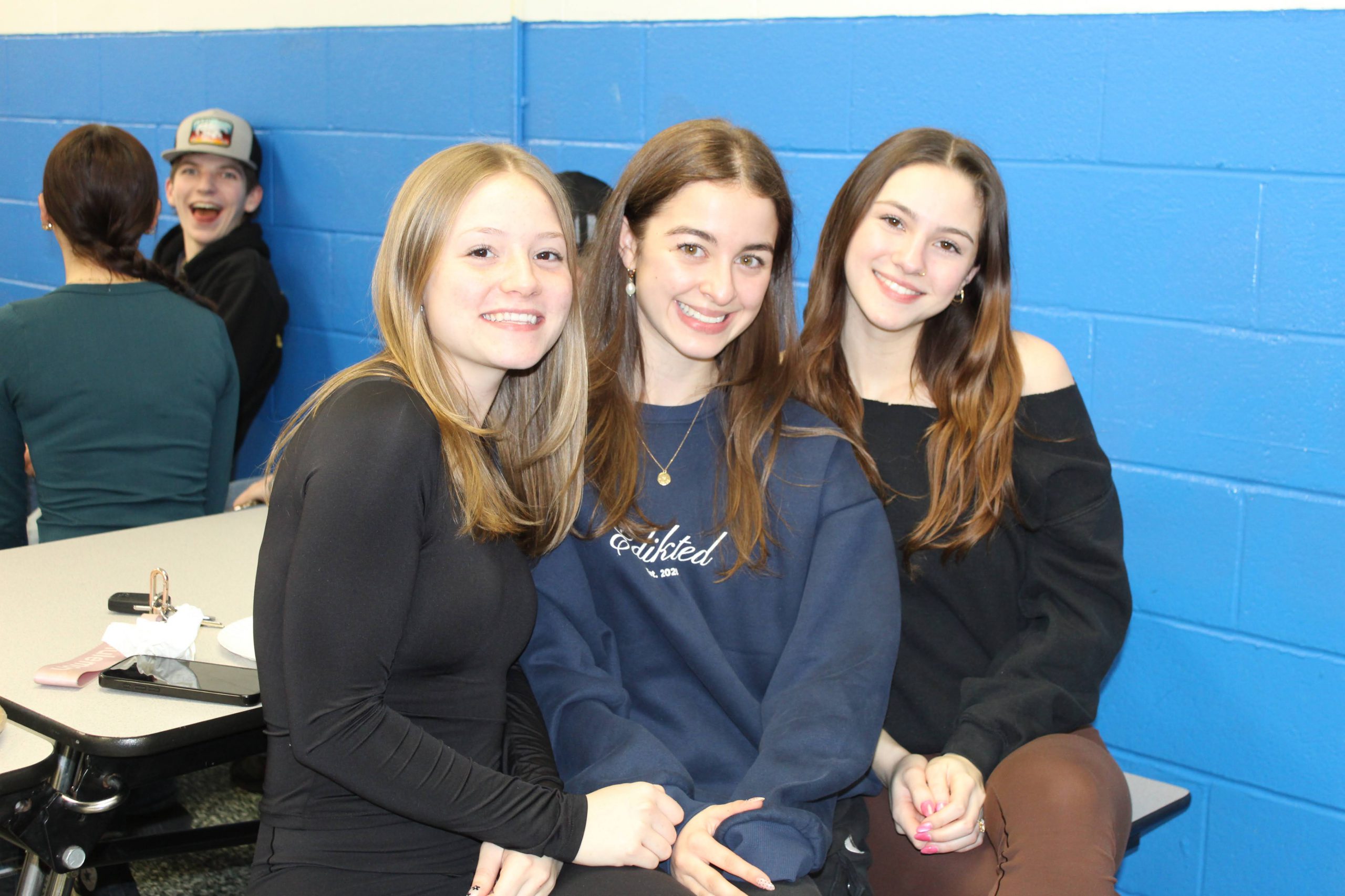 Three girls sit together in the cafeteria. A student in the background is smiling at the camera as well. 