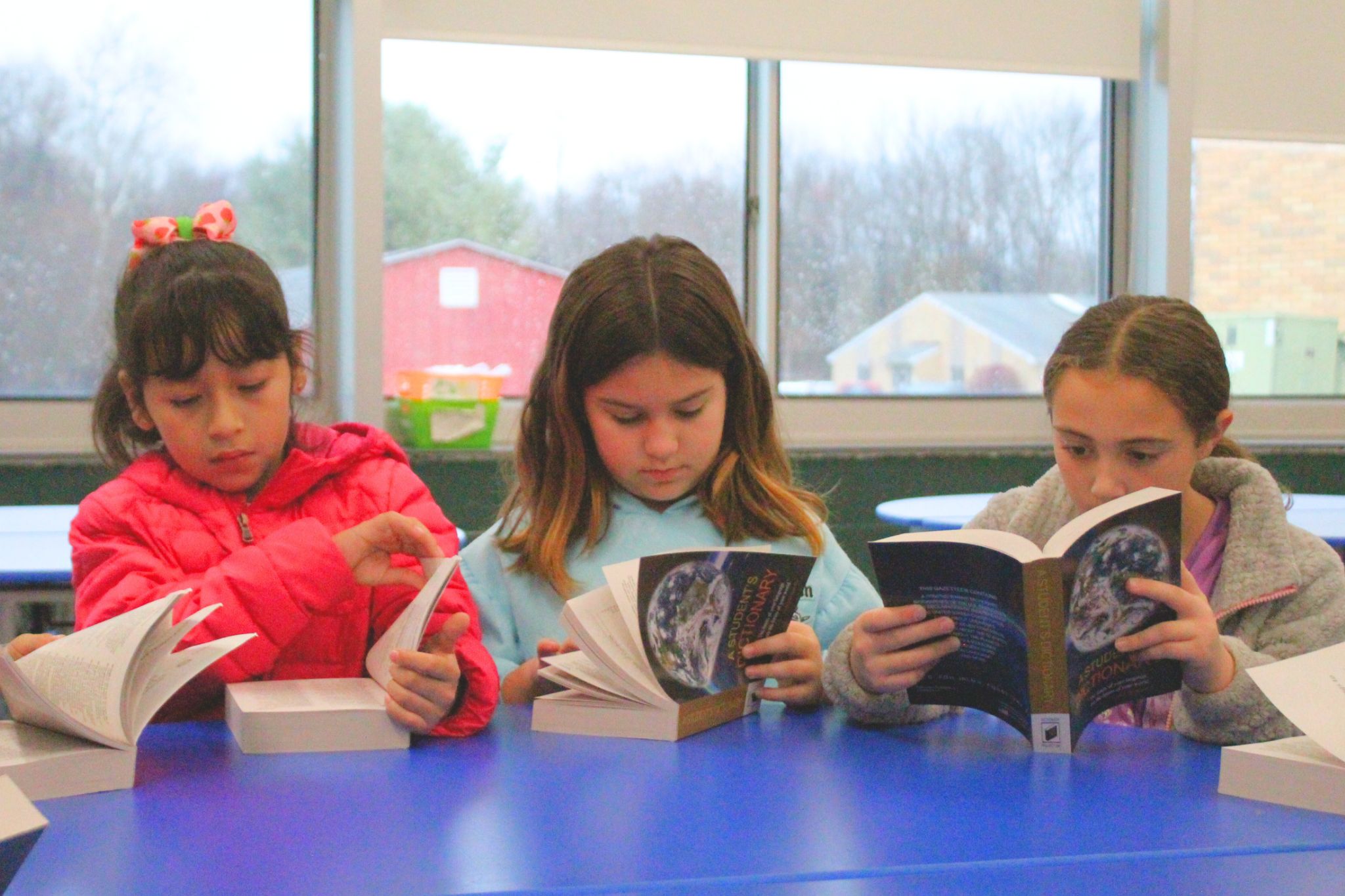 Three students reading a book in the cafeteria sitting next to each other. 