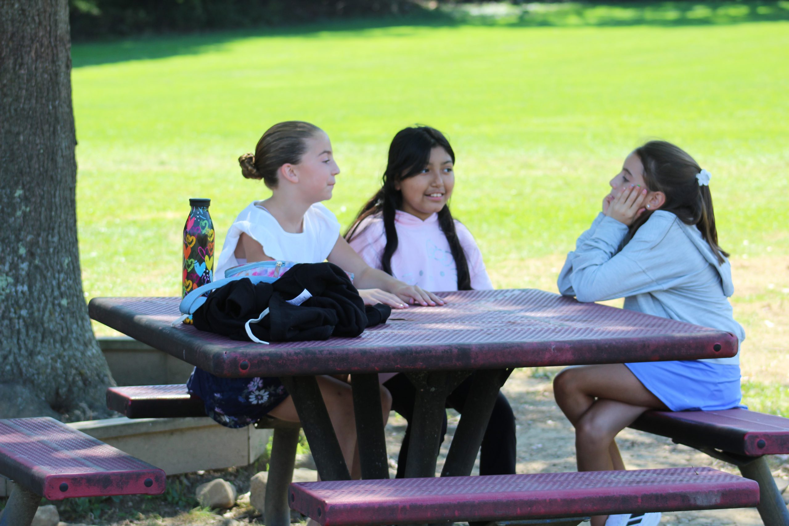 Three girls sit at a table outside talking on a sunny day. 