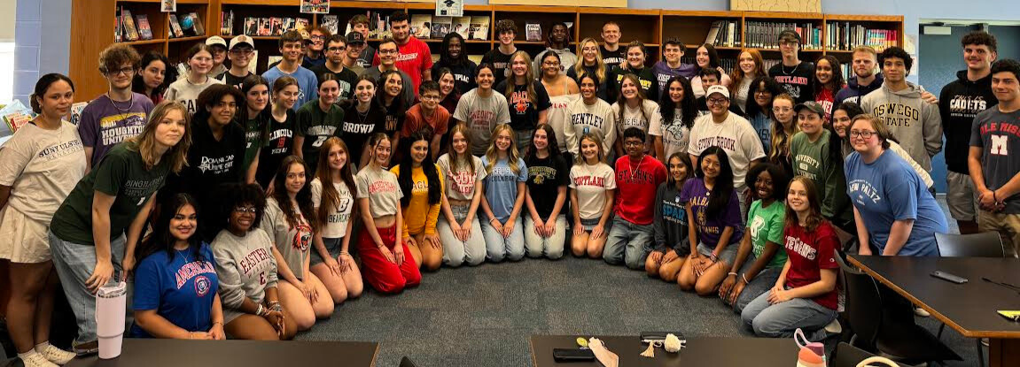 Group photo of VCHS seniors gathered in the library wearing shirts with the names of the colleges they plan to attend in the fall on them.