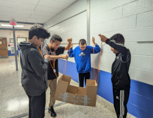 Students hold up cardboard with strings in the hallway as they work on a teambuilding activity. 