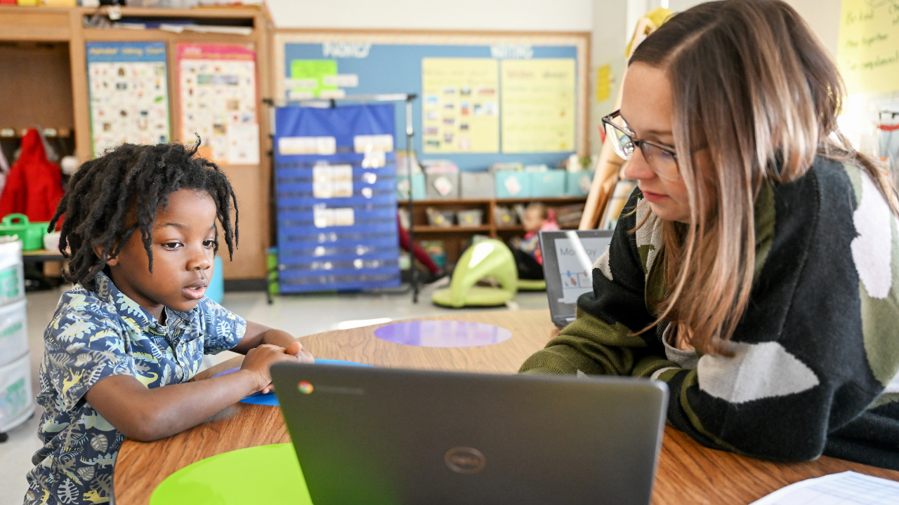 A student looks at a computer with a teacher nearby.
