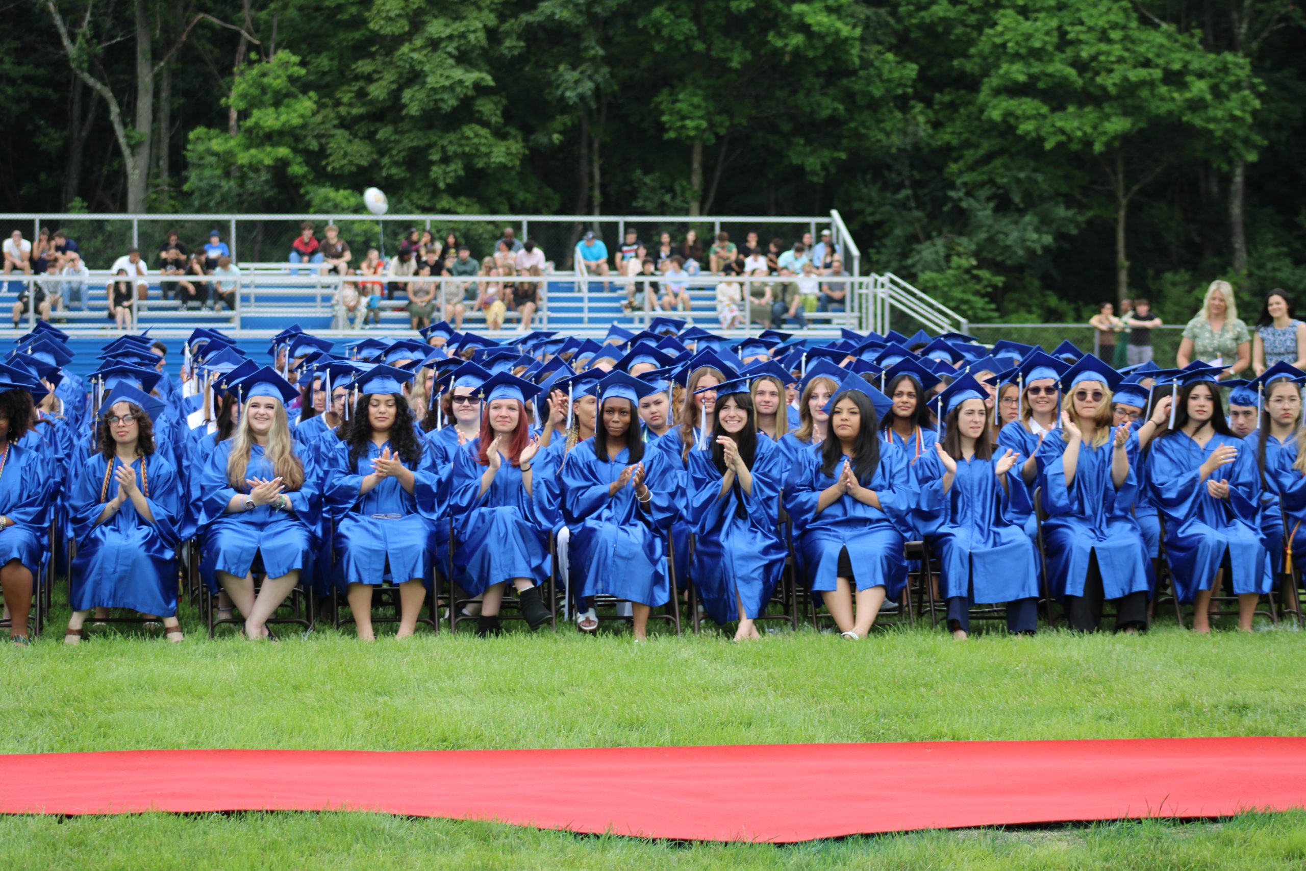 Students are sitting and clapping on the field during graduation in their cap and gown. 