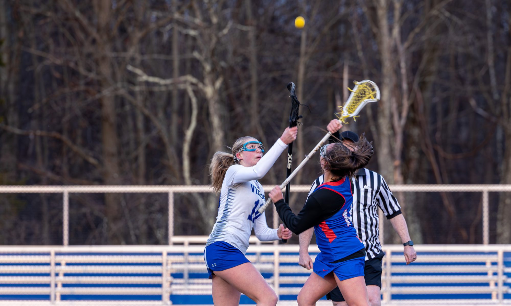 Two girls playing lacrosse on the field with the ball in the air above them.