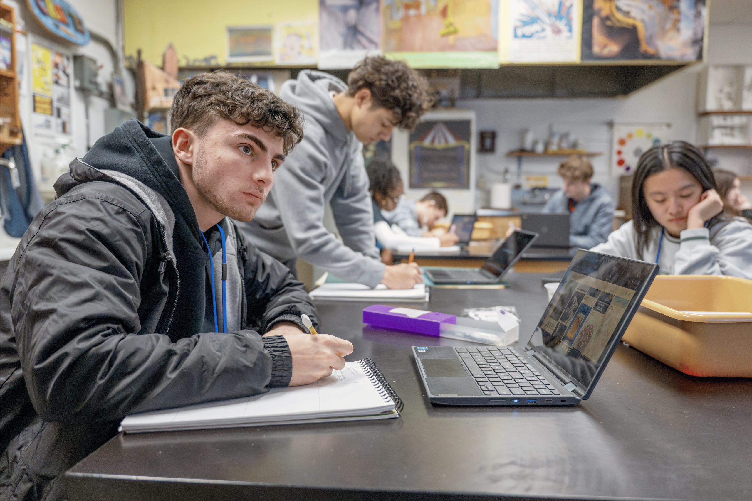 Boy sitting at a table writing in a notebook with a computer open in front of him. There is another student sitting at his table.