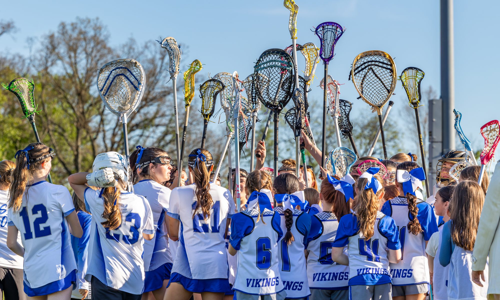 VC girls varsity lacrosse team holding their sticks in the air together.