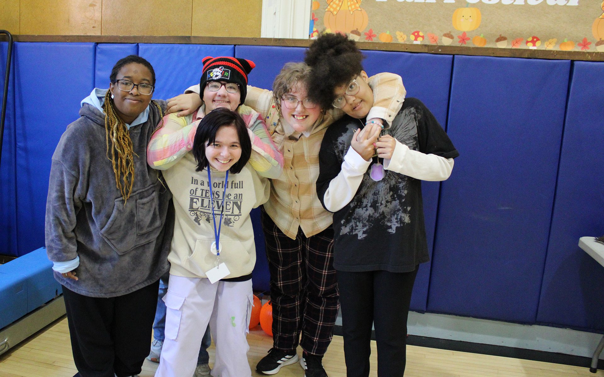 Five people wrap their arms around each other as they pose for a photo in front of an ALC Fall Fest sign